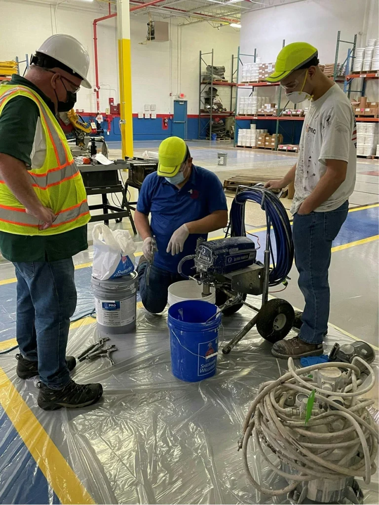 Three workers mix coating material beside spray equipment and buckets on plastic sheeting; a supervisor watches.