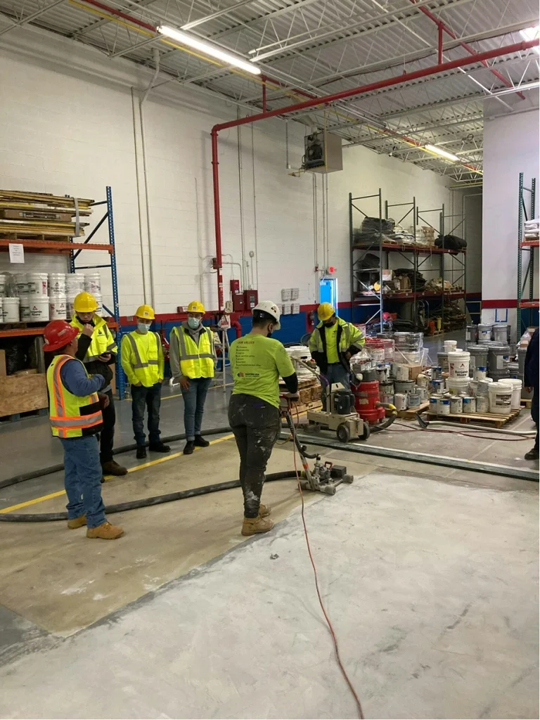 Warehouse demo: a worker grinds a concrete floor while a group in hard hats and vests observes from a safe distance.