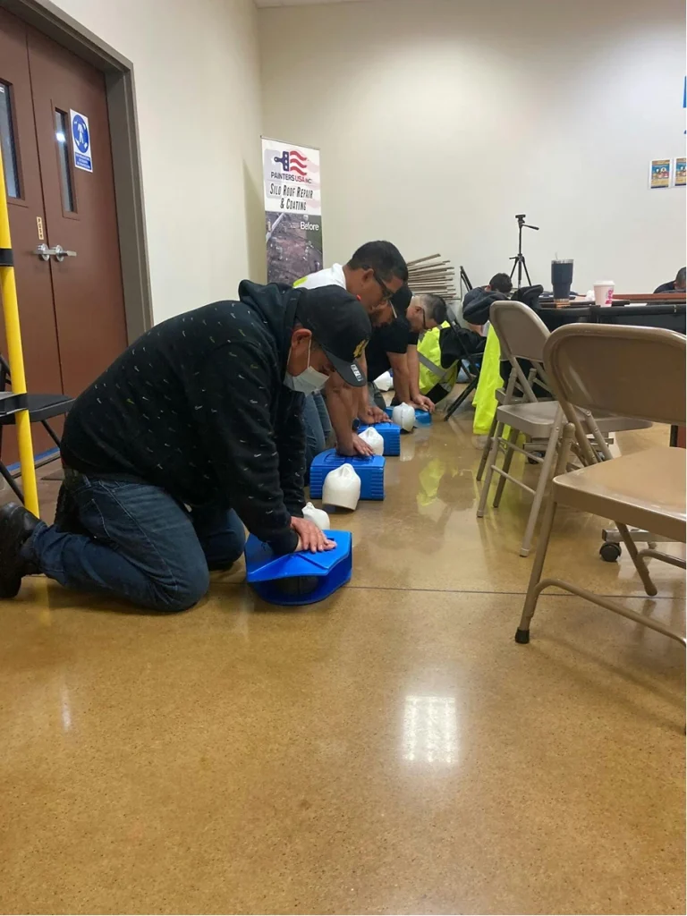 Trainees kneel in a row practicing CPR chest compressions on training mannequins along a classroom wall.