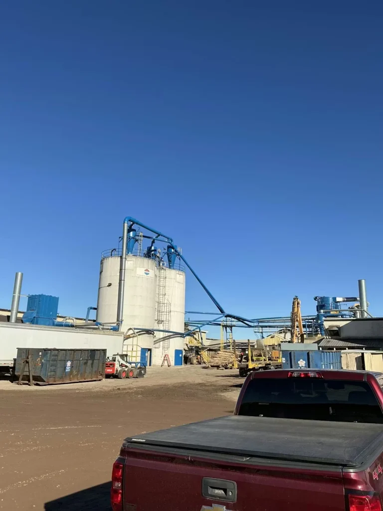 Industrial facility yard with large cylindrical silos and blue piping under a clear blue sky; a red pickup truck is parked in the foreground.