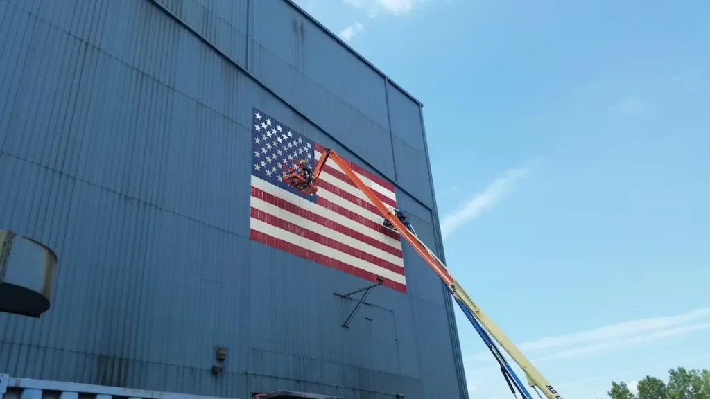 Workers in a boom lift repaint a large American flag mural on the side of a tall industrial building under a bright blue sky.
