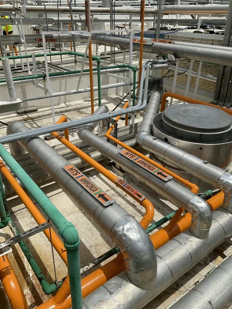 Network of industrial pipes and conduits on a rooftop, including orange lines and large insulated pipes labeled “HTS AMMONIA,” beside a round vent fan housing.