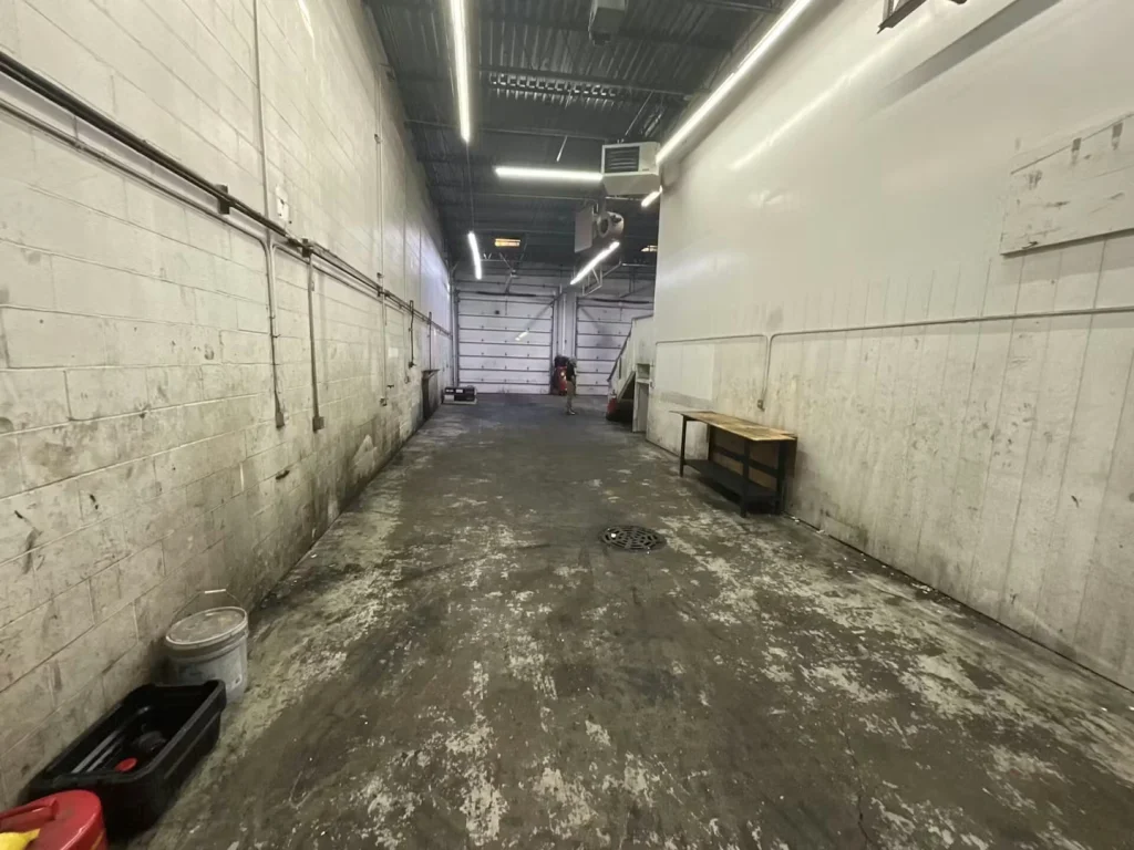 Long, narrow industrial bay with white block walls, a stained concrete floor, and a roll-up door at the far end.