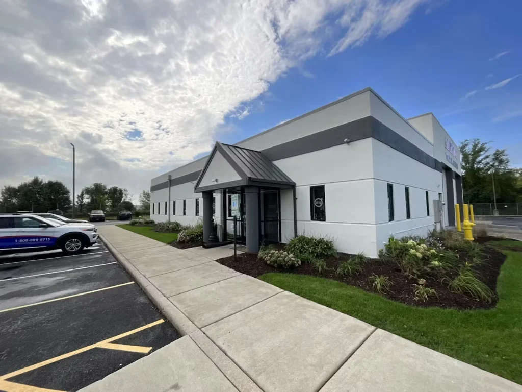 Exterior of a white commercial building with a small covered entrance, sidewalk, and parking lot under a partly cloudy sky.