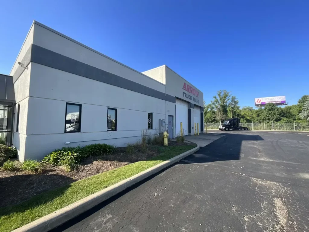 Exterior of a white commercial building with “Arrow Truck Sales” signage, blacktop driveway, and a semi-truck in the lot under a clear blue sky.