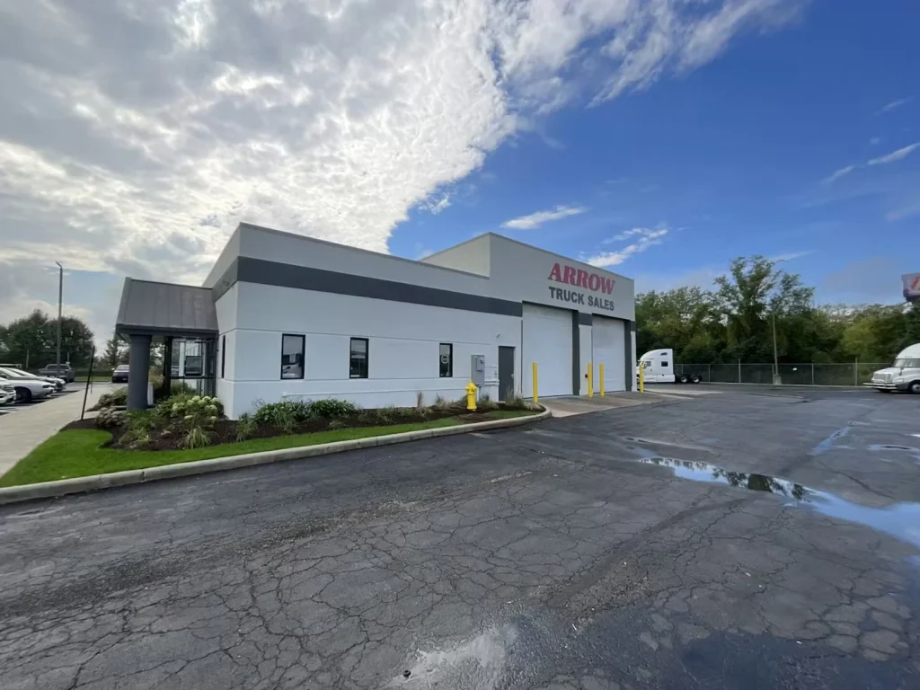 Wider view of the “Arrow Truck Sales” building and parking lot after rain, with puddles on the asphalt and trucks parked near service bay doors.