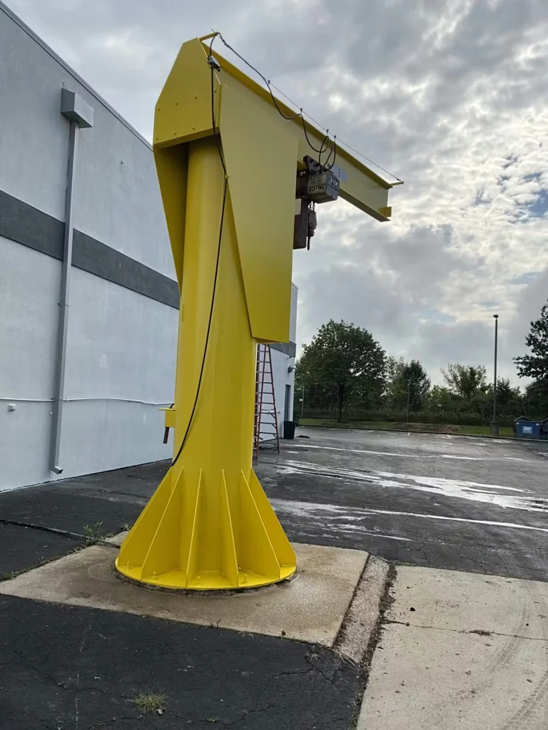 Close-up of a yellow jib crane base and boom with hanging hoist hardware, set next to a warehouse and parking lot.