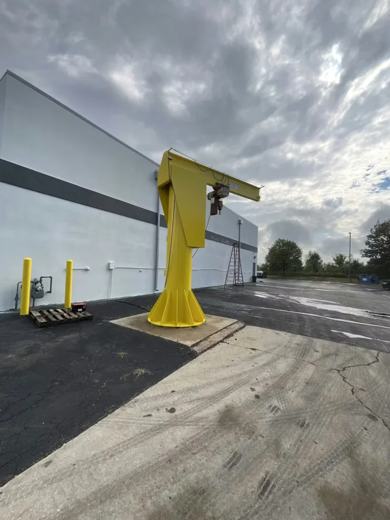Yellow freestanding jib crane with a hoist stands on a concrete pad beside a warehouse wall, with a ladder nearby under cloudy skies.