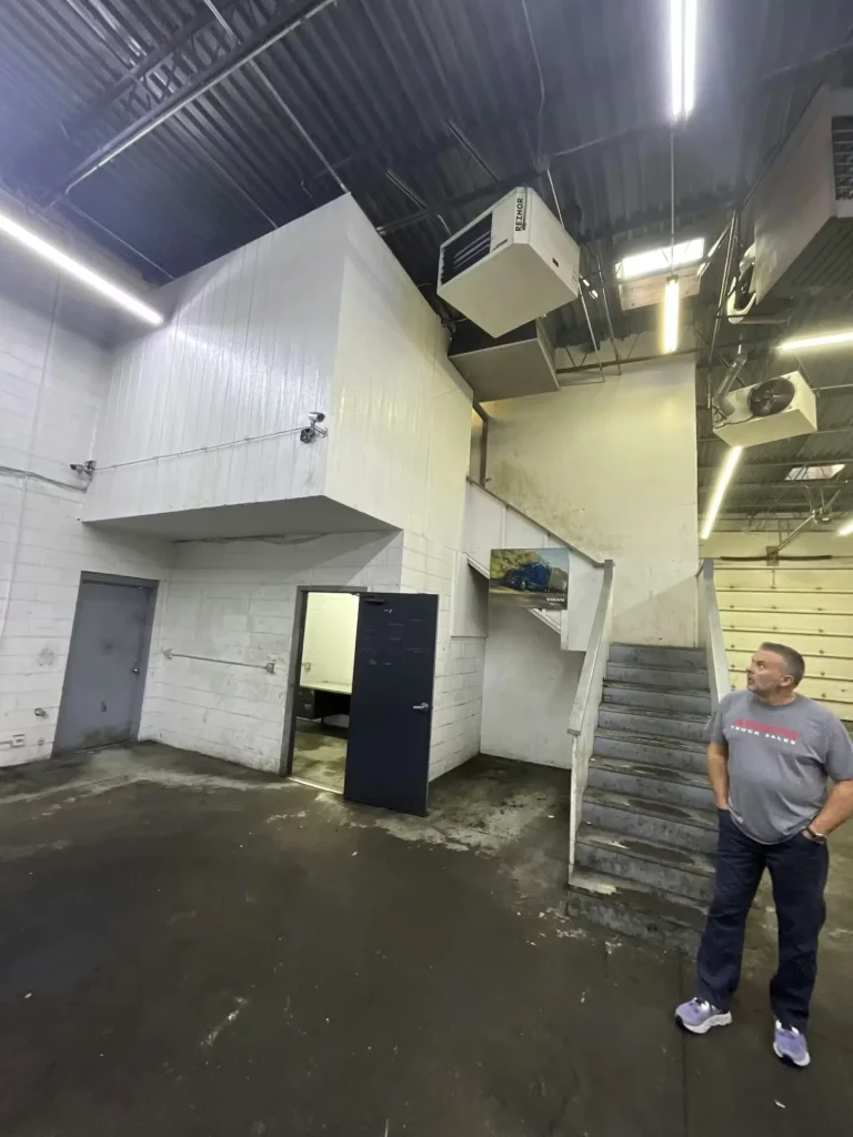 Warehouse interior with a mezzanine and stairs; a person stands near doorways under exposed ceiling equipment.