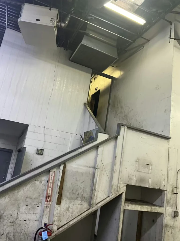 Close view of worn warehouse stairs and mezzanine structure with scuffed, dirty white surfaces and exposed ducting above.