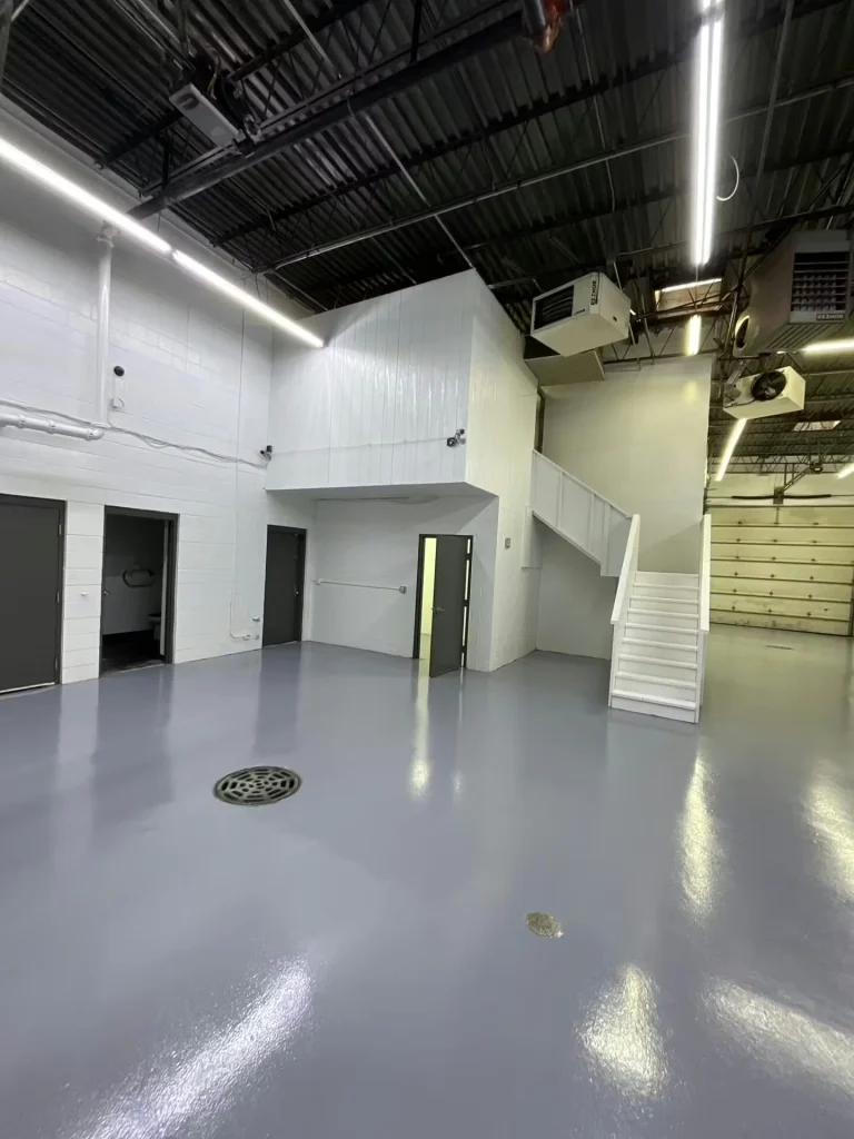 Wide warehouse space with bright white walls, doors along one side, and a glossy gray floor beneath exposed ceiling equipment and stairs.