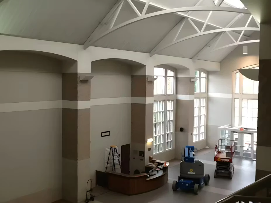 Large interior atrium with high vaulted truss ceiling and tall windows; a reception desk, ladders, and two lift machines sit on the floor during painting or maintenance.