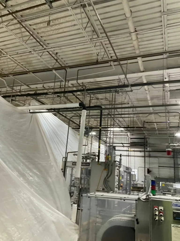 Interior of an industrial facility with an exposed metal ceiling and overhead piping; large equipment below is partially covered with clear plastic sheeting.