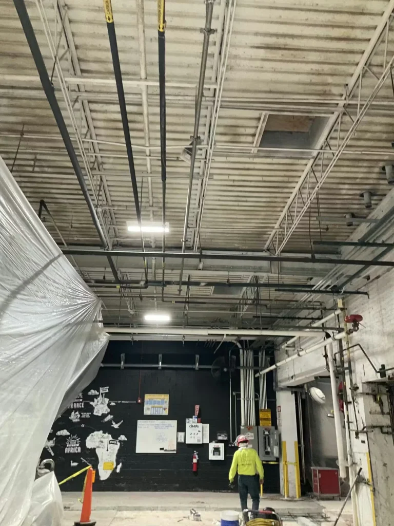 High-ceiling industrial room with exposed beams and hanging pipes; a worker in a high-visibility shirt and hard hat stands near a dark wall with a mural, with plastic sheeting draped along the left side.