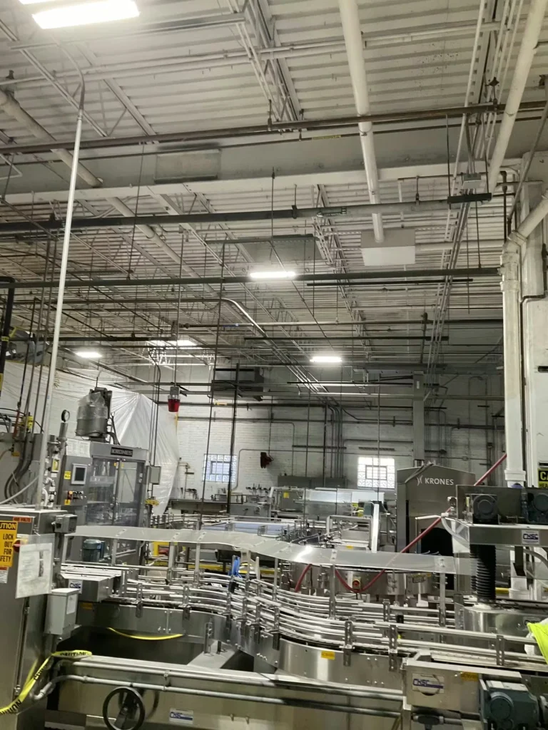 Brewery production area with stainless-steel conveyors and machinery under a high, exposed ceiling with bright overhead lights and utility lines.