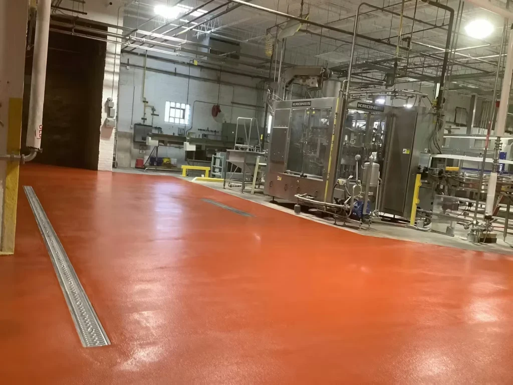 Wide view of a brewery floor coated in a glossy orange-red finish, with a long floor drain channel on the left and stainless equipment in the background.