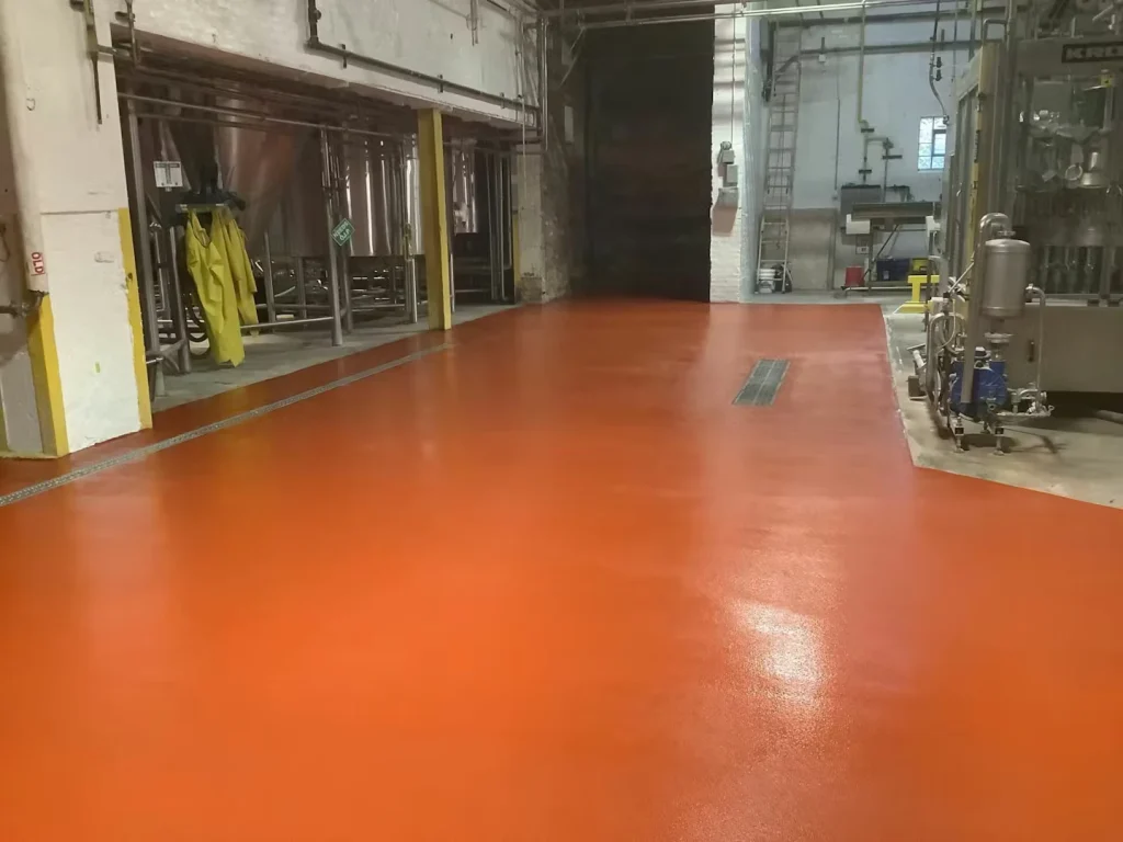 Orange-red coated brewery floor leading toward an open doorway; stainless tanks and piping line the sides, and a rectangular floor drain grate sits near the center.