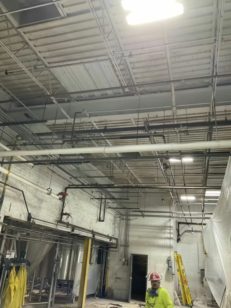 Industrial interior with exposed ceiling and suspended piping; a worker in a high-visibility shirt and hard hat stands near a ladder and plastic sheeting along the right side.