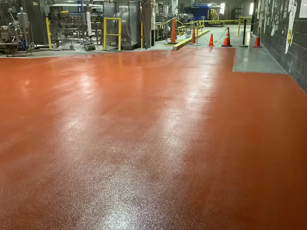 Large orange-red coated floor area sectioned off with caution tape and orange traffic cones, with stainless brewery equipment visible behind the taped boundary.