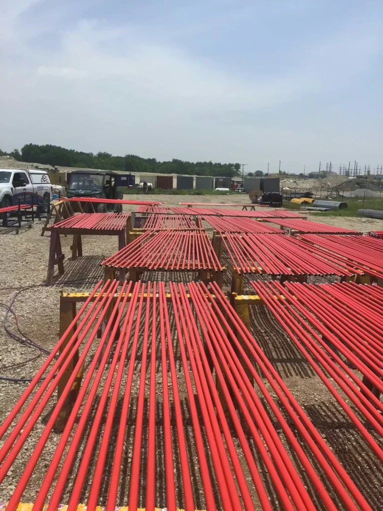 Outdoor work yard with many red-coated pipes laid out on sawhorses for drying, stretching into the distance under a bright sky.