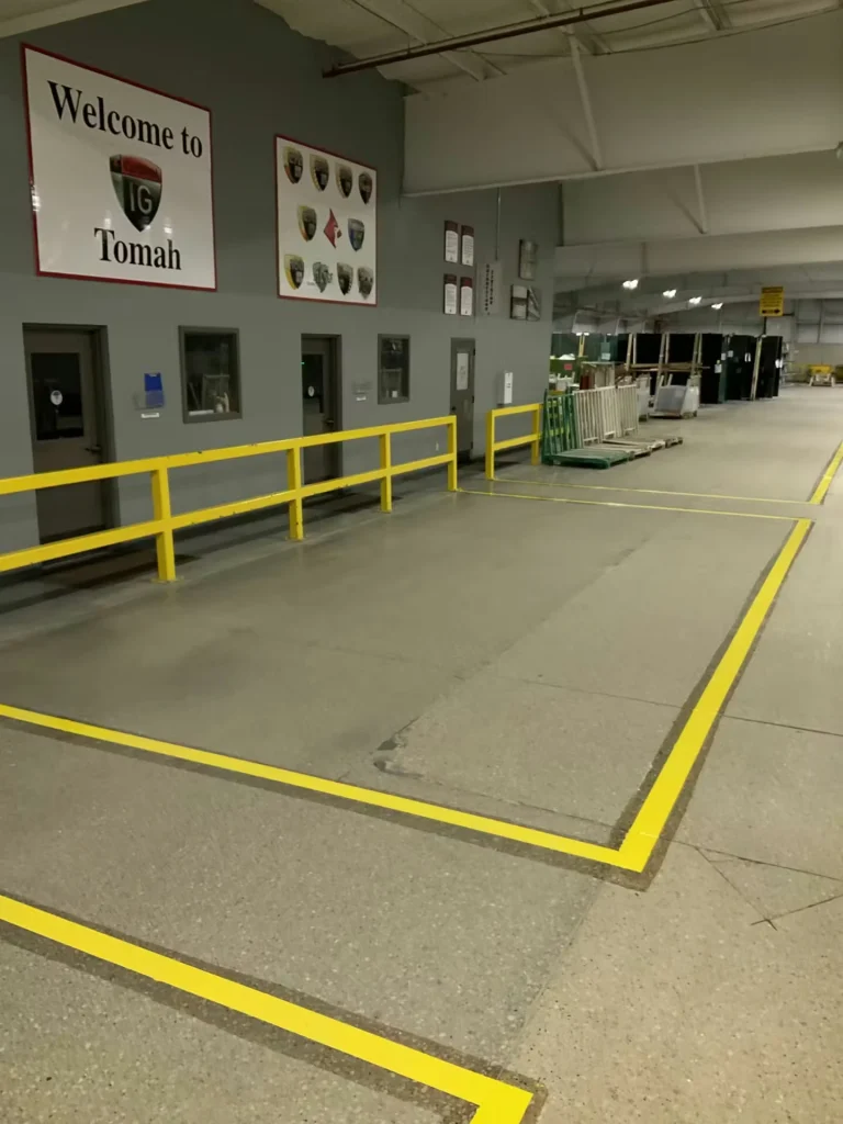 Industrial facility hallway with yellow safety railings and bright yellow floor markings; a wall sign reads “Welcome to Tomah,” with doors and windows along the left side and a large open work area extending to the right.
