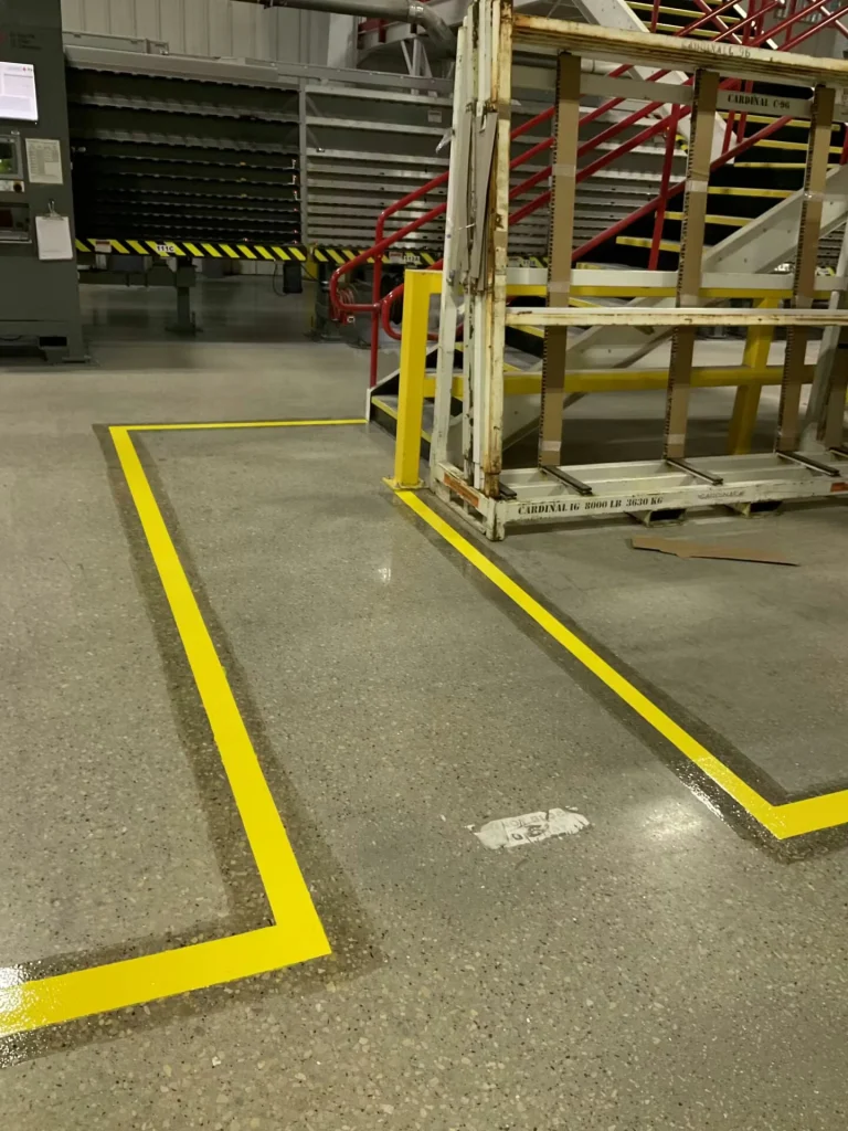 Wide factory aisle with racks of large glass panes on pallets to both sides; a forklift is parked in the middle distance under high warehouse lighting.