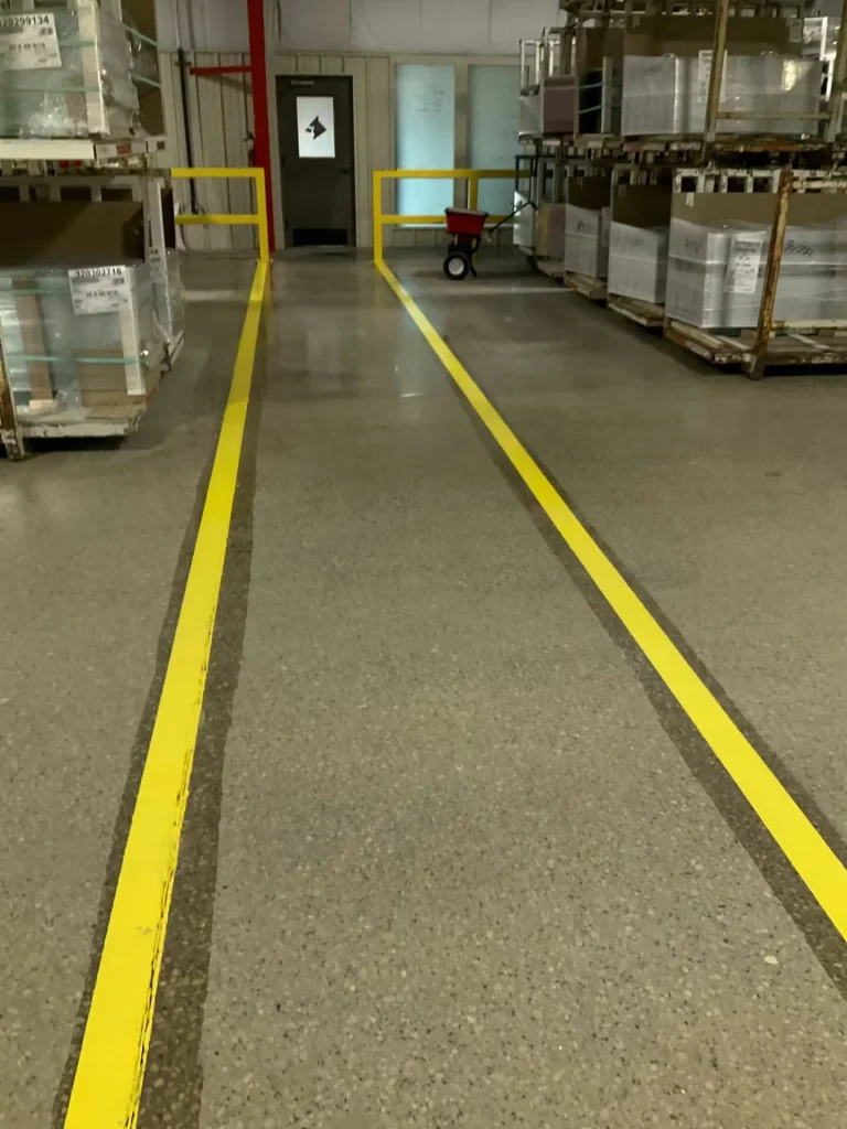 Factory floor with a yellow-lined pedestrian lane beside a corrugated wall; a small sign with an arrow points “OFFICE,” and glass-transport carts are visible ahead.