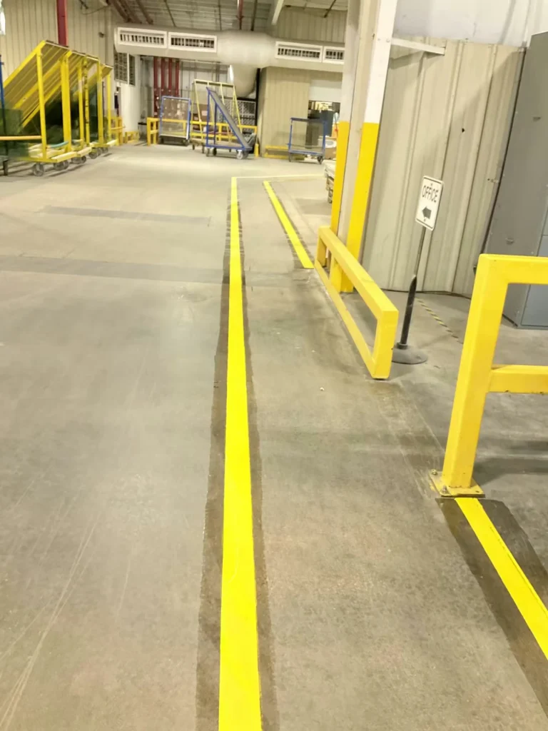 Concrete factory floor showing faint, worn yellow lane lines running down a long aisle; shelving and equipment appear far in the background.