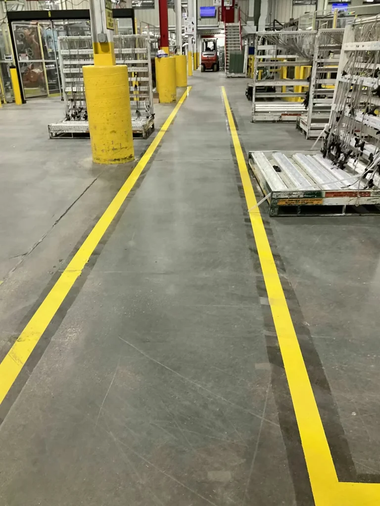 Straight pedestrian walkway marked by two bright yellow lines through a warehouse area; large yellow bollards and metal racks line the path, with a forklift visible ahead.