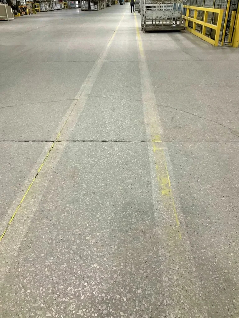 Similar warehouse walkway view with fresh bright yellow lane lines and an “OFFICE” arrow sign on the right; glass carts and equipment sit in the distance.