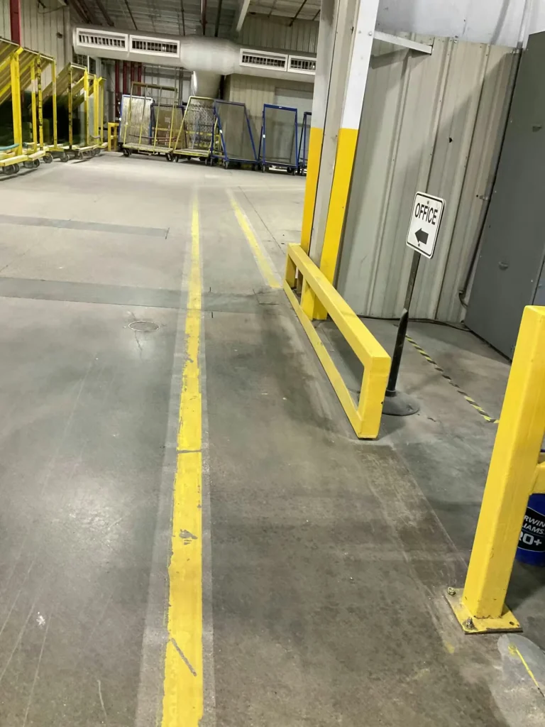 Warehouse corridor between pallet racks, with two parallel bright yellow floor lines guiding toward a door; a red wheelbarrow sits near the doorway.