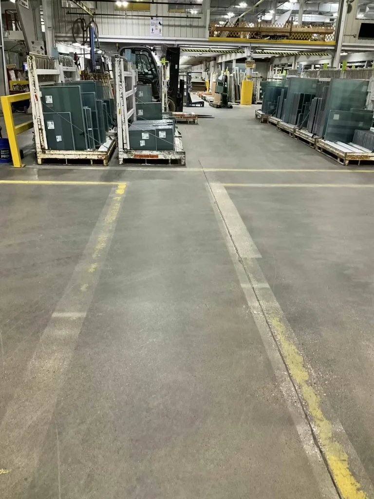 Concrete factory floor with freshly painted bright yellow rectangles outlining staging areas around equipment; a metal rack stands near red stairs and industrial machinery in the background.
