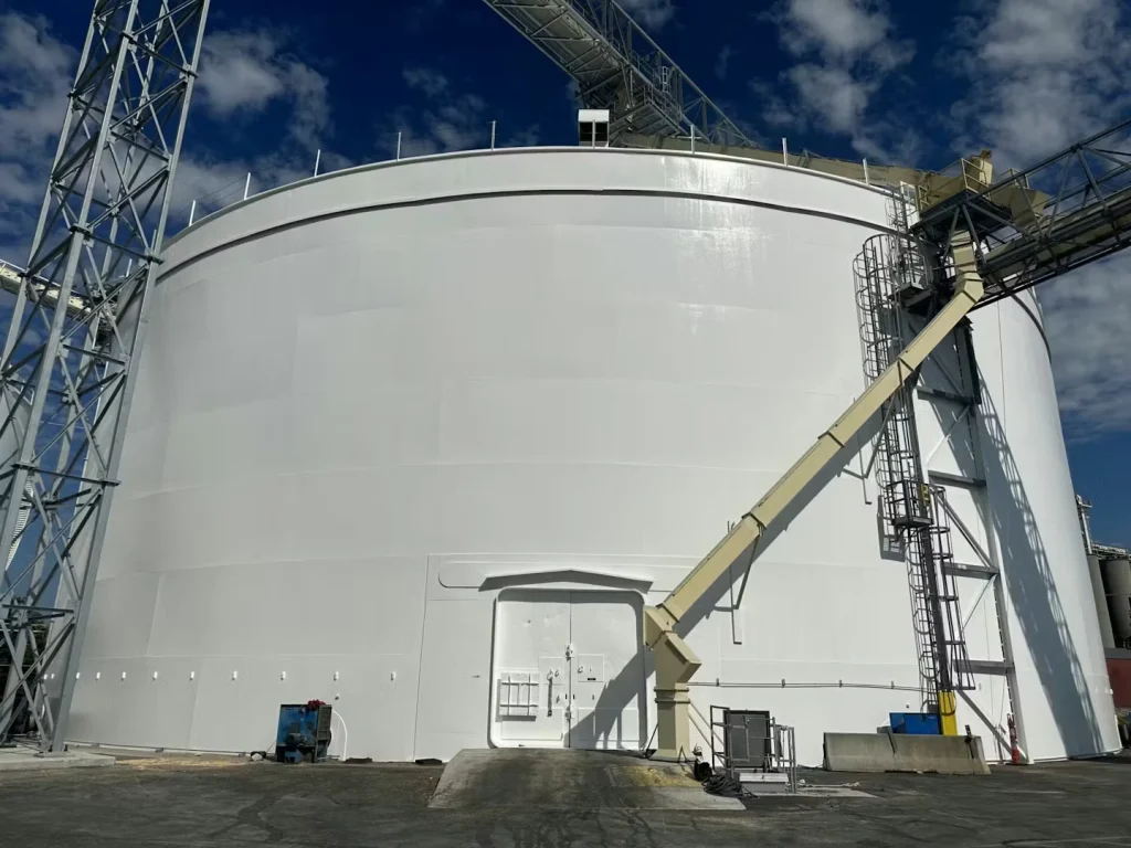 Close view of a massive white cylindrical storage tank under a blue sky with scattered clouds; metal catwalks and conveyor structures cross overhead, with a vertical ladder and platform attached to the tank’s right side.