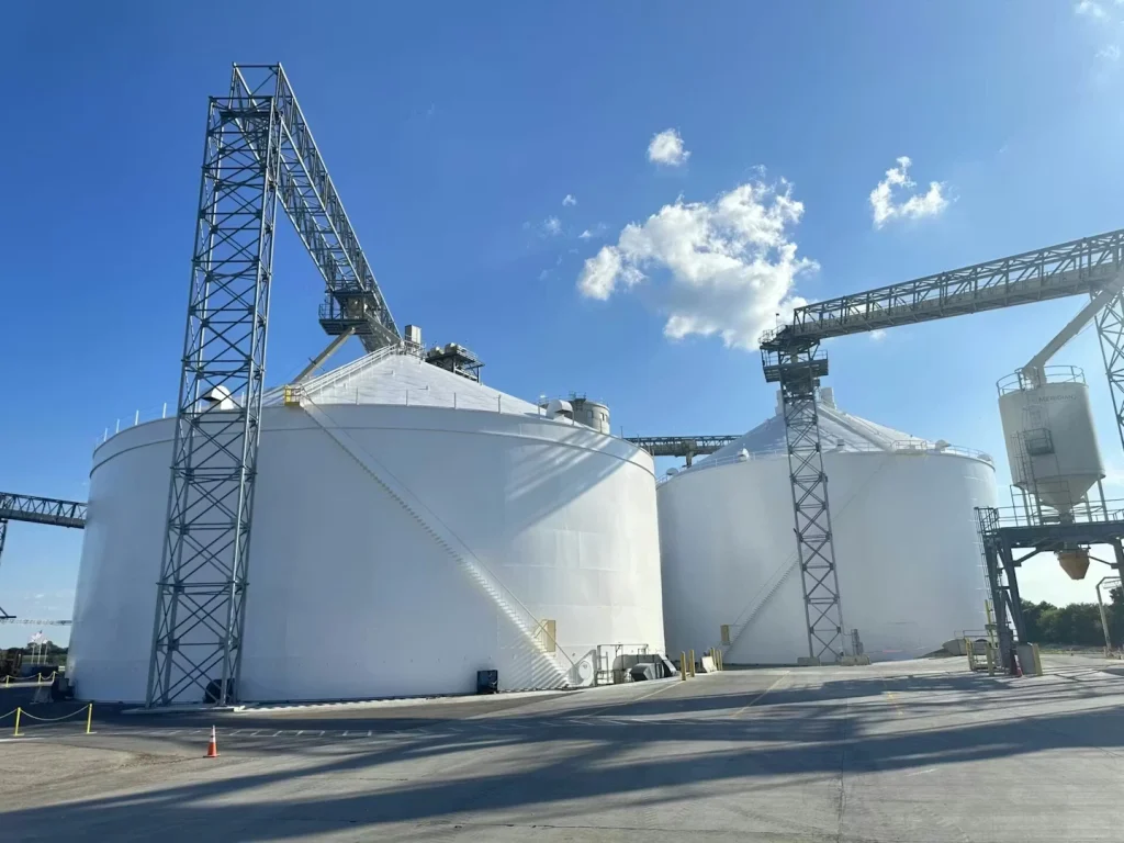 Wide outdoor view of multiple large white storage tanks with conical roofs; tall steel conveyor towers and catwalks span between tanks under a bright blue sky, with a paved yard and traffic cone in the foreground.