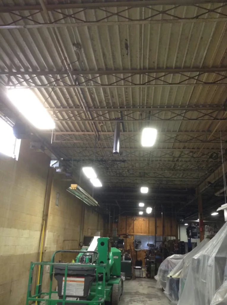 Tall industrial bay with an exposed metal roof and bright rectangular overhead lights; a green boom lift sits on the floor below, and machinery and equipment are partially covered with clear plastic along the right side.