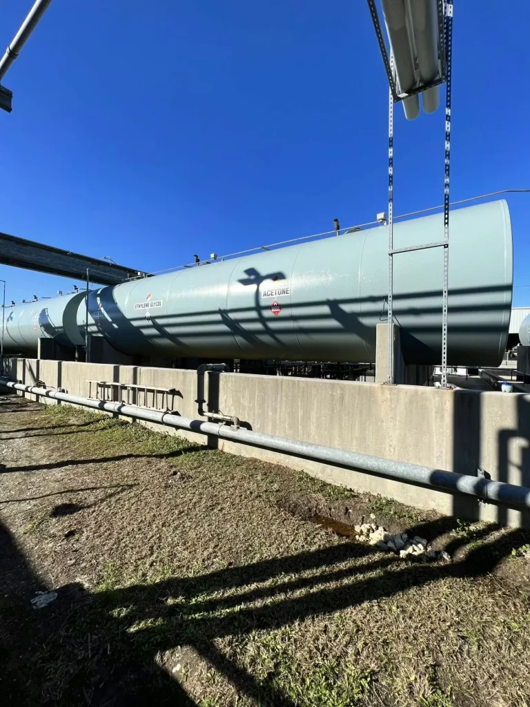 Two long horizontal storage tanks at an industrial facility, one labeled “Methanol,” with piping racks and valves on the left and shallow standing water reflecting the tanks on the concrete pad.