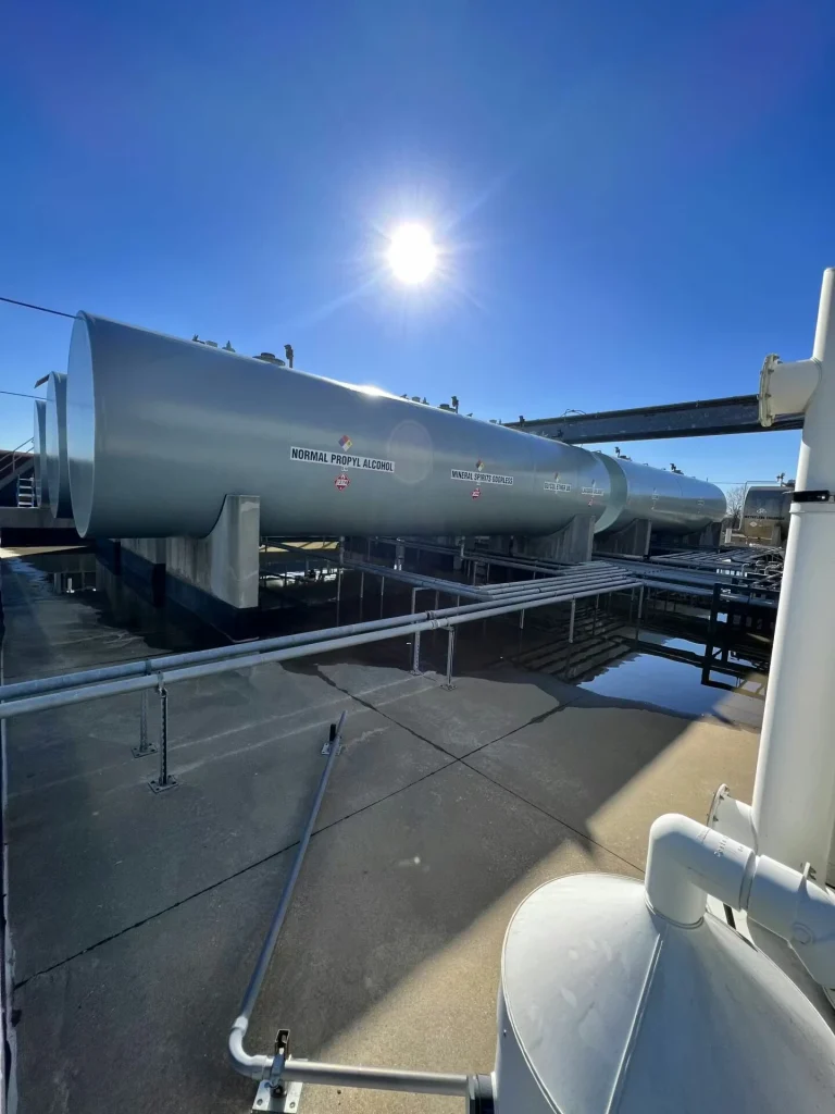 Row of horizontal chemical storage tanks with hazard placards; the nearest tank is labeled “Normal Propyl Alcohol,” with the sun high overhead and catwalk shadows crossing the tanks and ground.