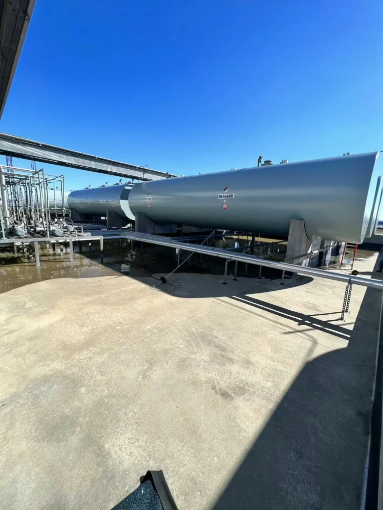 Horizontal storage tank labeled “Acetone” behind a concrete containment wall; overhead pipe supports cast strong shadows across the tank, with grass and a pipe run in the foreground under a bright blue sky.