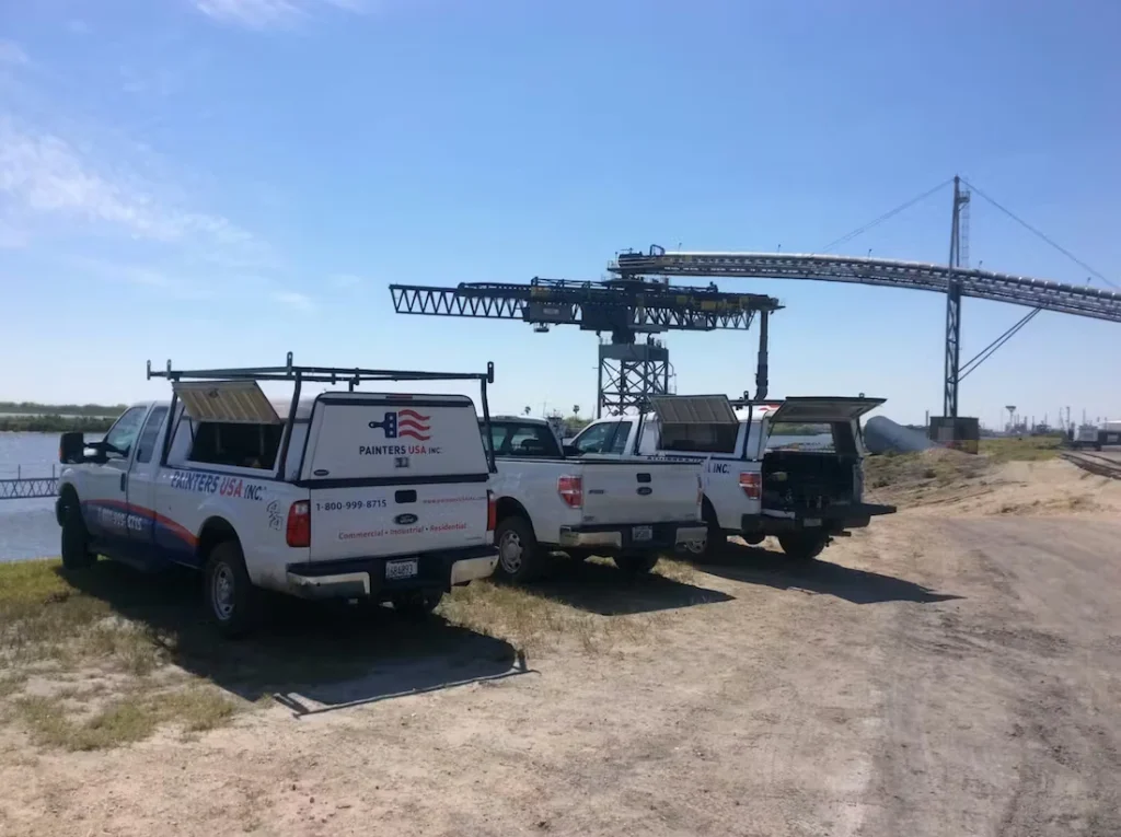 Three work pickup trucks parked on a dirt road beside a waterway, with toolboxes and ladders on the beds; a large industrial ship loader/conveyor structure rises in the background under a clear blue sky.