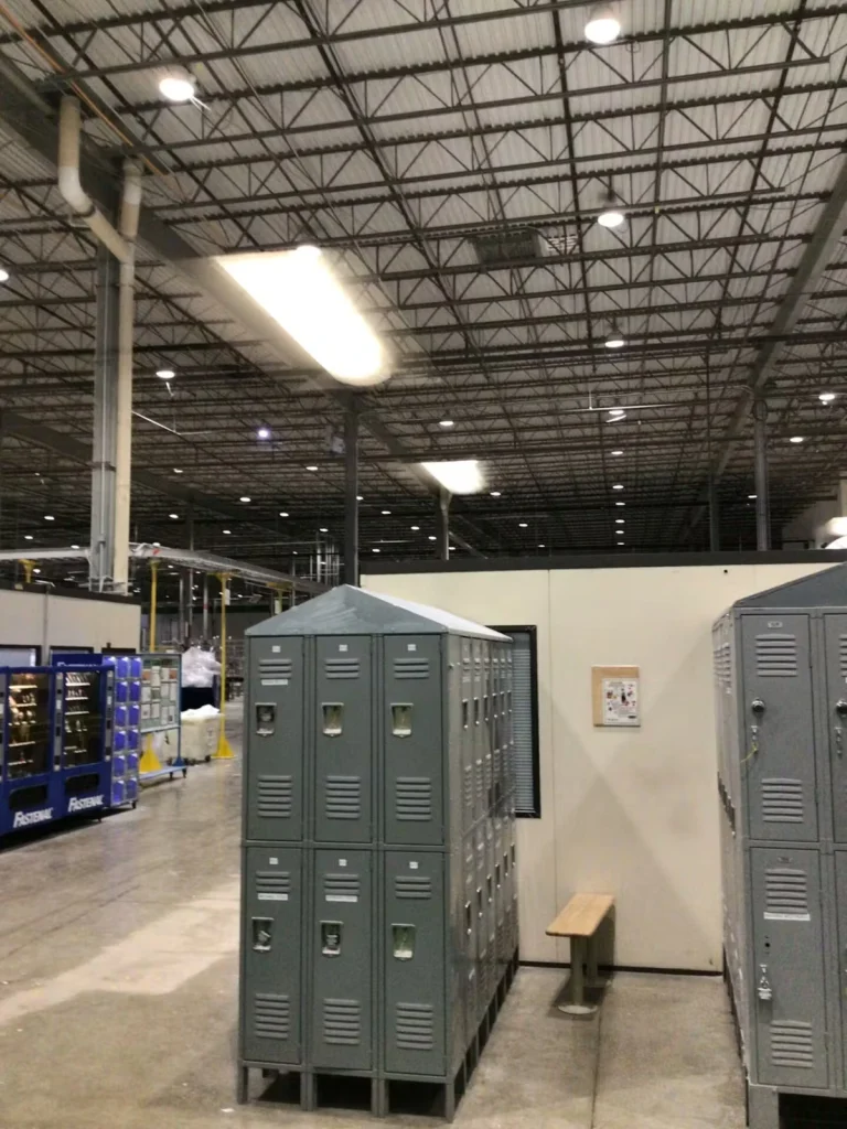 Inside a large industrial warehouse, rows of gray metal lockers sit beside a small white office enclosure with a bench, under a high truss ceiling with bright overhead lights and equipment racks in the background.