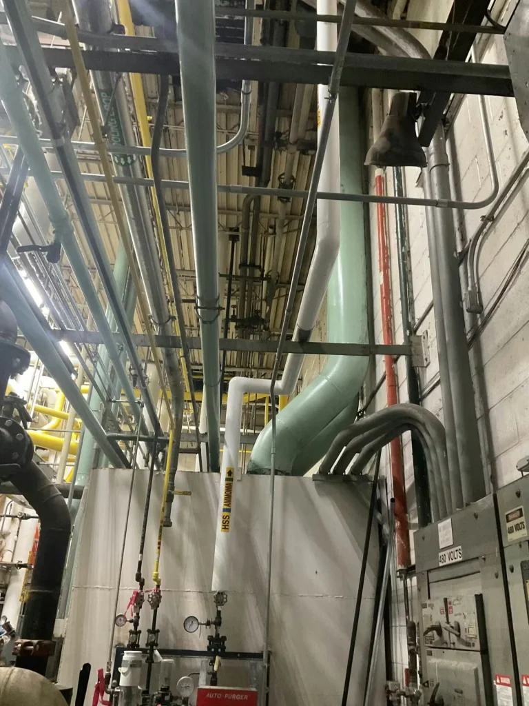 Interior industrial mechanical room with dense overhead piping and conduit; large pale-green ductwork bends down along a white wall, with gauges/valves and a metal equipment enclosure below, and an electrical panel labeled “480 VOLTS” at right.