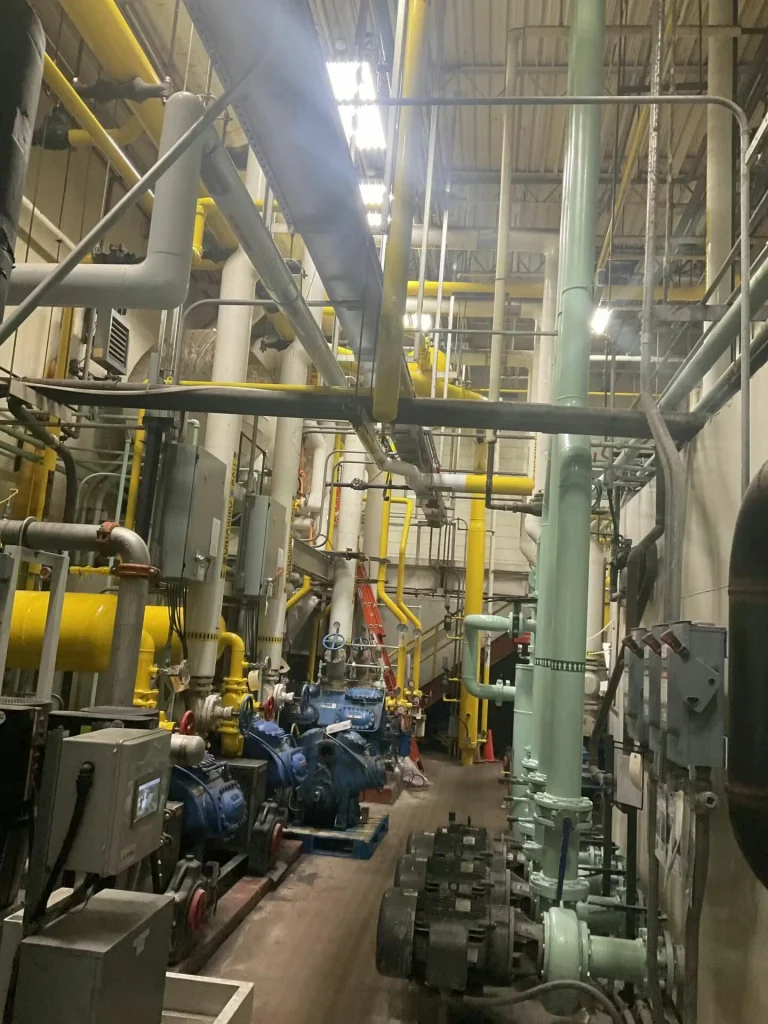 Close view of several tall mint-green vertical pipes with angled elbows near the top, smaller dark pipes crossing behind them, and electrical panels mounted on the wall.