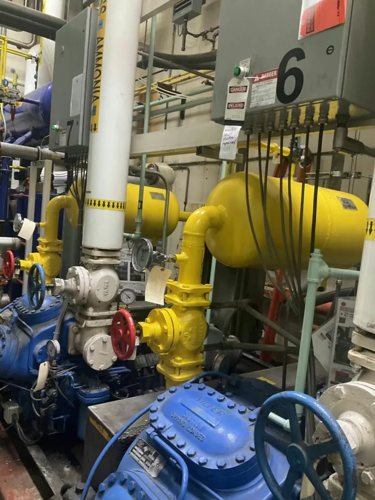 Close view of industrial equipment: a large blue machine with gauges and a yellow valve/pipe assembly, surrounded by structural steel framing and many intersecting pipes.