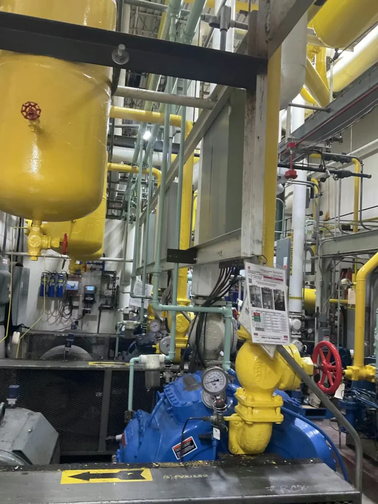 Wide view down an industrial mechanical room with dense overhead piping, yellow-painted lines and supports, large blue pump/compressor units on the floor, and catwalk/handrail framing in the foreground.