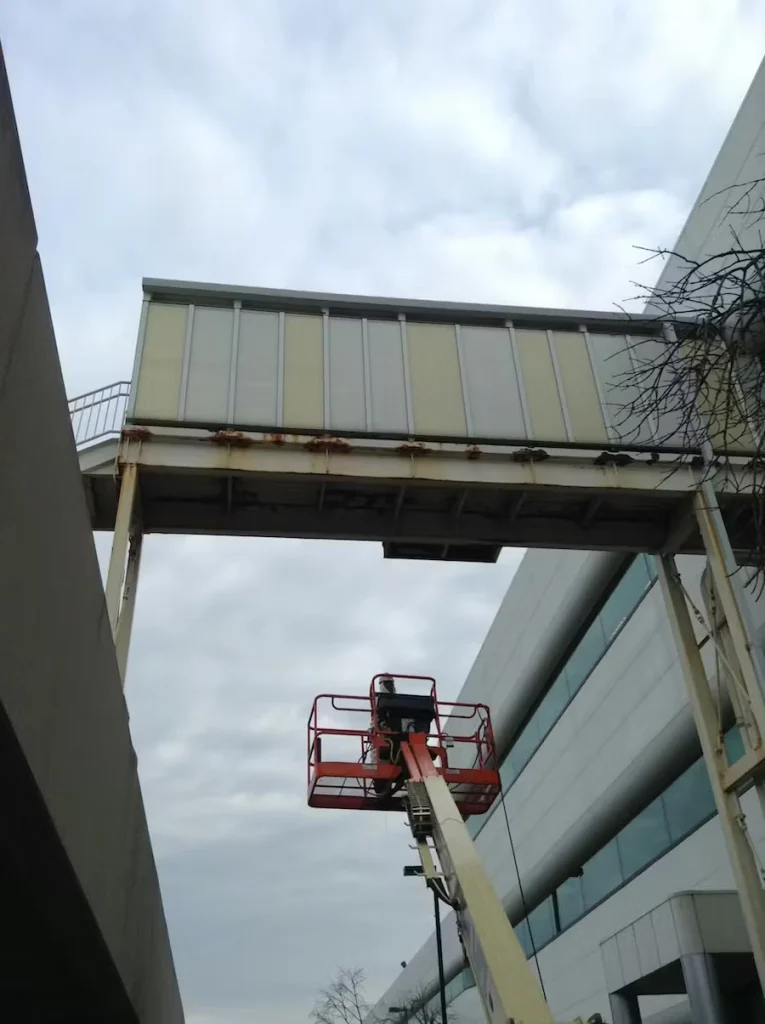 A worker stands in a red boom lift basket raised between two buildings, reaching toward the underside of an elevated enclosed walkway/skybridge with visible rust staining along the metal support beam under an overcast sky.