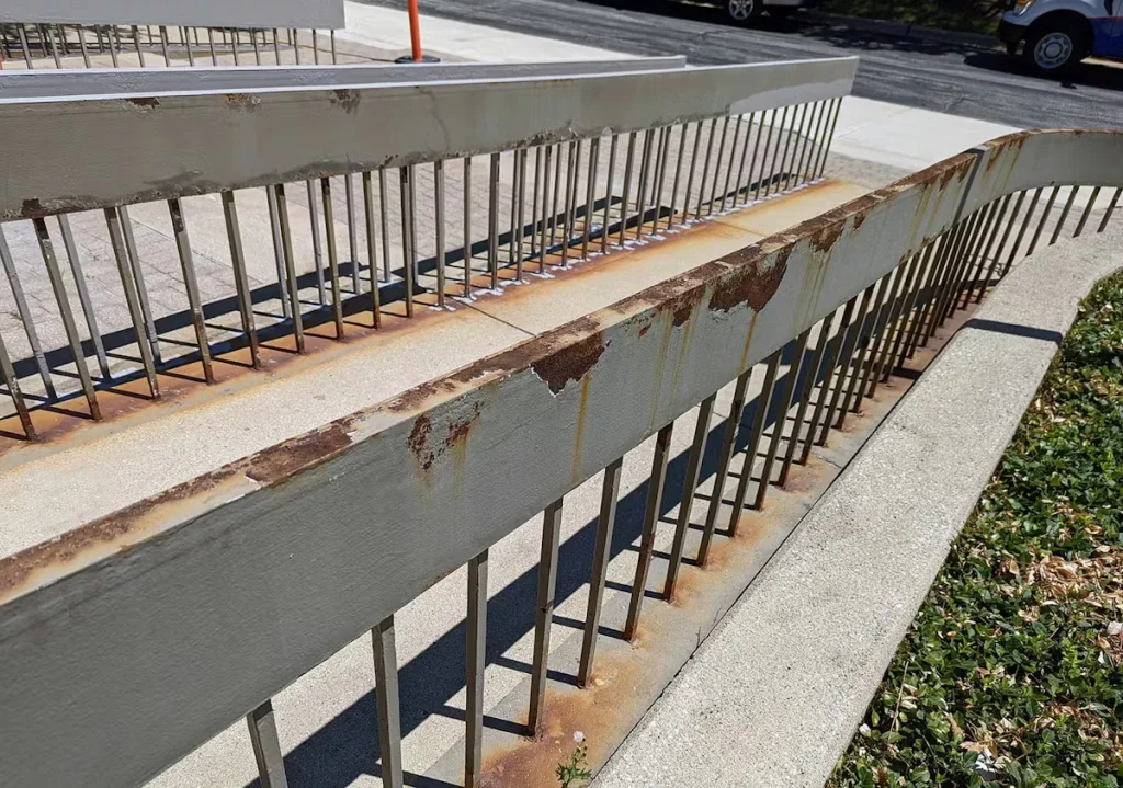Close-up of a gray-painted metal handrail with areas of chipped paint and rust along the top edge; the walkway and rail balusters run diagonally into the background.
