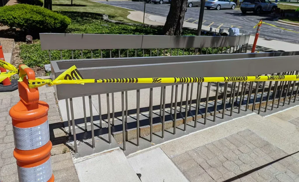 Side view of the gray handrail showing rust staining and patchy paint along the lower edge; white filler or sealant is visible at the base where vertical bars meet the concrete.