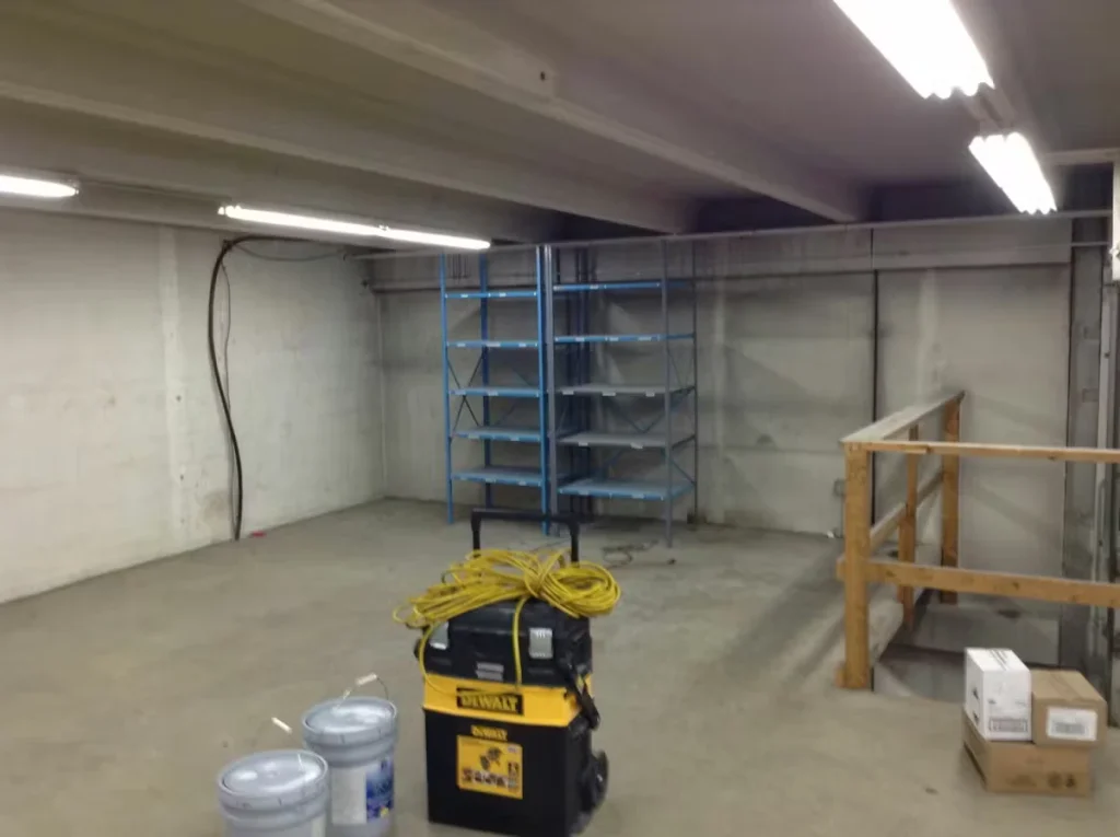 Empty concrete room with fluorescent strip lights on the ceiling; a yellow-and-black shop vacuum with coiled extension cord sits in the foreground next to two paint buckets, with blue metal shelving against the back wall and a wooden guardrail on the right.