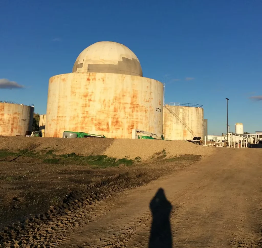 Large cylindrical industrial storage tank with a domed top, stained with vertical rust streaks, seen from a dirt access road at a plant site under a clear blue sky; smaller tanks and piping are in the background, and a person’s shadow falls in the foreground.