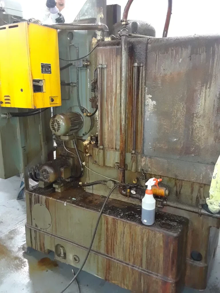 Grimy industrial machine with a bright yellow control box, exposed motors and pipes, and heavy oil/rust streaks running down the metal panels; a spray bottle sits on the lower ledge and the wet floor shows pooled residue, with a worker partially visible at the top edge.